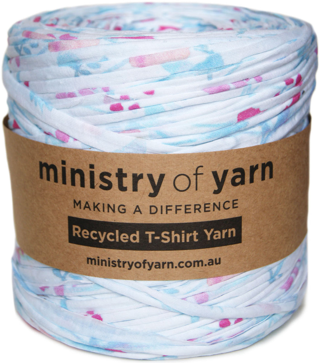 A spool of Ministry of Yarn recycled T-shirt yarn with a brown paper label, featuring a white base with light blue and pink printed patterns.