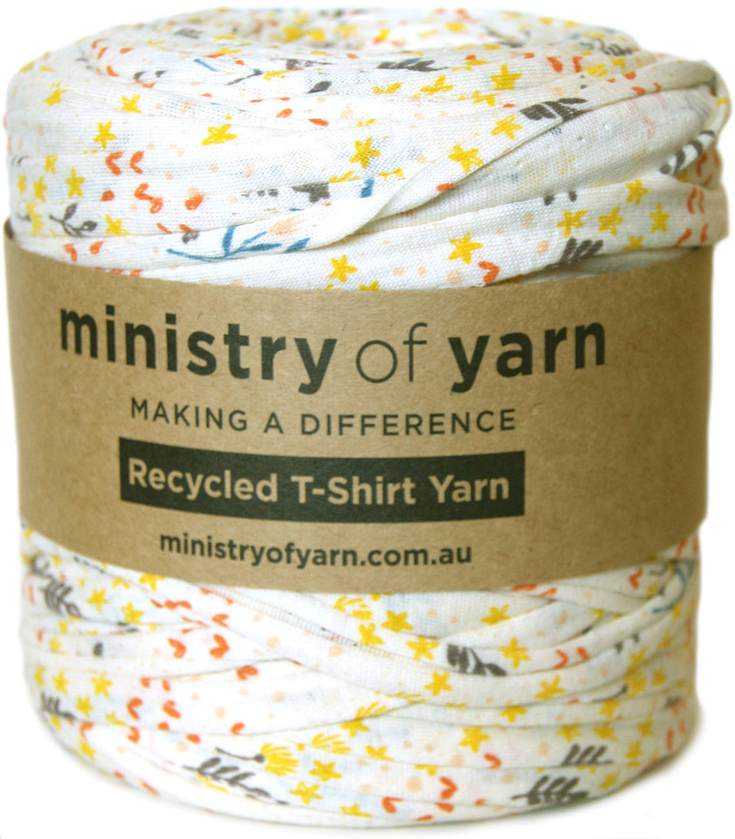 A spool of Ministry of Yarn recycled T-shirt yarn with a brown paper label, featuring a white fabric base with a colorful print of yellow stars, orange leaves, and gray leaves.