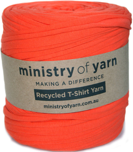 A bright orange-red roll of yarn with a paper label reading 'ministry of yarn' and 'Recycled T-Shirt Yarn' on a white background.