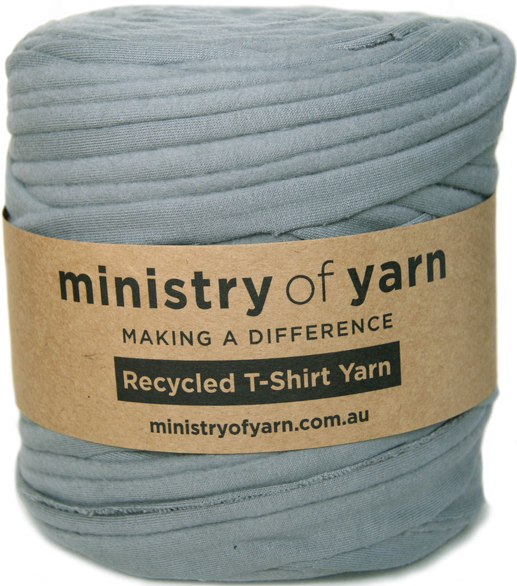 A roll of dusty blue-gray recycled T-shirt yarn wrapped with a brown kraft paper label that reads 'ministry of yarn' and 'Recycled T-Shirt Yarn'.