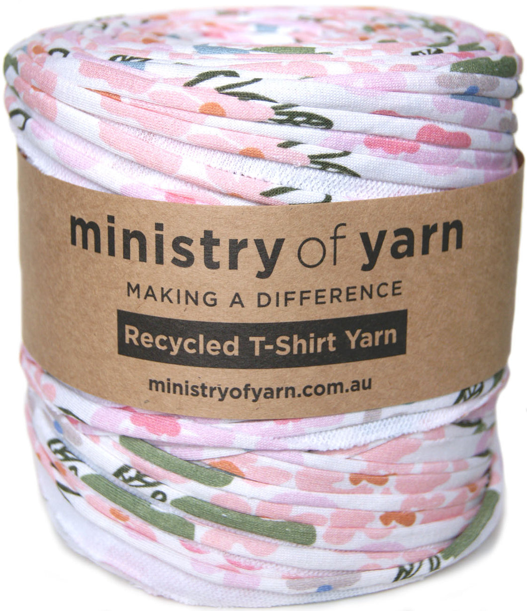 A spool of Ministry of Yarn recycled T-shirt yarn wrapped in a brown paper band, featuring a pink, green, and blue abstract pattern on a white background.
